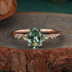 May include: A rose gold engagement ring featuring an oval green gemstone surrounded by smaller green gemstones.