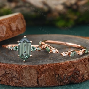 May include: A rose gold engagement ring set with a green octagon-shaped gemstone and a matching rose gold band with green and white gemstones. The ring is on a wooden surface.