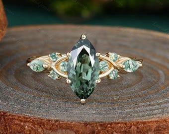May include: A gold ring featuring a large, marquise-cut green gemstone as the centerpiece. The ring is adorned with smaller, light green gemstones and intricate gold detailing. The ring is set on a wooden surface.