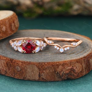 May include: A rose gold engagement ring set. The main ring has a central, round, red gemstone, encircled by white gemstones. The accompanying band is curved, with small white gemstones. The rings are on a wooden surface.
