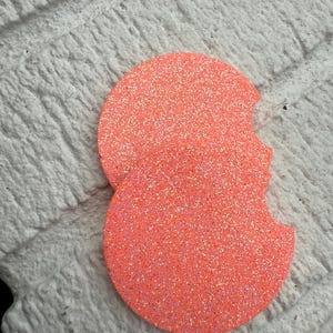 May include: Two coral-colored car coasters with a glittery finish. The coasters are round with a small notch on one side. They are displayed on a white, textured surface.