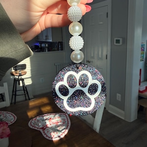 May include: A round, glittery black and purple paw print charm with a white paw print design. The charm is attached to a beaded strand of white pearls and textured beads.