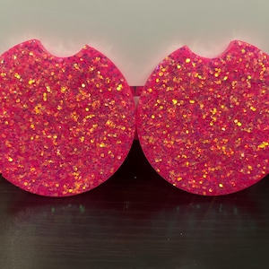 May include: Two pink round glitter coasters with a cut-out for a cup. The coasters are covered in iridescent glitter.