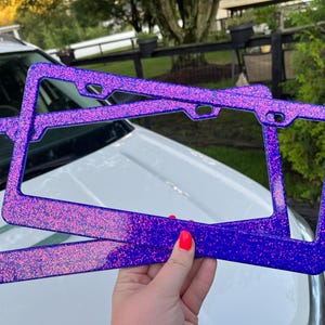 May include: Two purple license plate frames with glitter. The frames are rectangular with rounded corners and holes for mounting. The glitter is a mix of pink and blue, creating a shimmering effect. The frames are held in front of a white car.