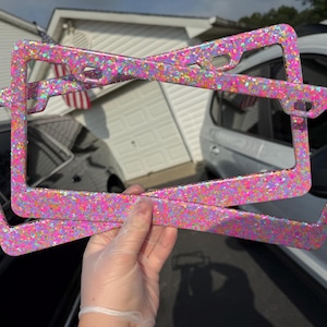 May include: Two glitter-covered license plate frames in shades of pink, purple, and orange are held up against a background of a house and cars. The frames are rectangular with rounded corners and openings for the plate and mounting hardware.