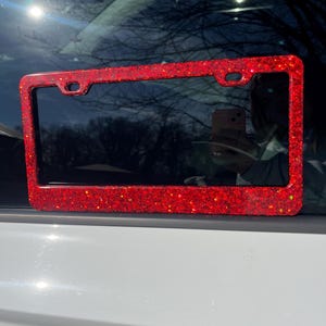 May include: Red glitter license plate frame. The frame is made of a shiny red glitter material and has a black metal border.