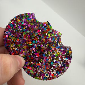 May include: A round car coaster with a cutout for easy removal. The coaster is covered in colorful glitter, including red, pink, green, blue, and gold. The glitter is embedded in a clear resin.