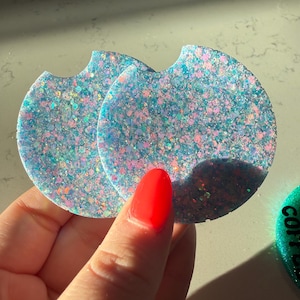 May include: Two round car coasters with a glittery, speckled design in shades of blue, pink, and iridescent colors. Each coaster has a curved notch on one side. The coasters are held by a hand with a red painted nail.