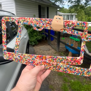 May include: Two license plate frames with a colorful sprinkle design are held up in front of a car. The frames are rectangular with rounded corners and a clear background. A small brown tag with a heart is attached to the top frame. The frames are held by a person's hand.