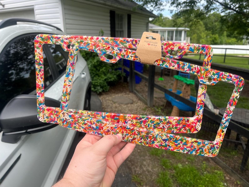 May include: Two license plate frames with a colorful sprinkle design are held up in front of a car. The frames are rectangular with rounded corners and a clear background. A small brown tag with a heart is attached to the top frame. The frames are held by a person's hand.