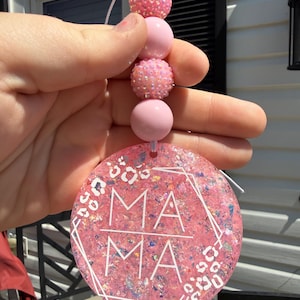 May include: A round pink resin ornament featuring a glittery finish, a white geometric frame, and a white leopard print pattern. The word MAMA is printed in white in the center. The ornament hangs from a string of four beads, alternating between smooth pink spheres and textured, iridescent pink spheres.