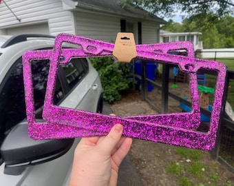 May include: Two bright pink glitter license plate frames are held up in front of a car. The frames are rectangular with rounded corners and have a sparkly finish. A small brown tag with a heart is attached to the frames.