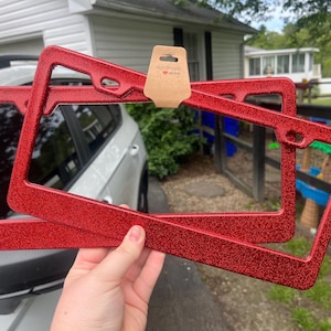 May include: Two red glitter license plate frames are held up in front of a car. The frames are rectangular with rounded corners and a textured, sparkly finish. A small tag with the words "handmade with love" is attached.