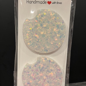 May include: Two handmade car coasters with a glittery, iridescent design. The coasters are white with a circular shape and a small notch on one side. The packaging reads "Car Coasters" and "Handmade with love."