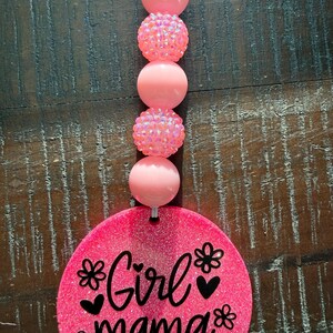 May include: A pink glittery car charm with the words "Girl mama" in black script, decorated with flower and heart accents. The charm is strung with pink beads, including some with a sparkly, textured finish.