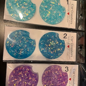 May include: Set of six car coasters in three colors: two light blue, two medium blue, and two purple. Each coaster is round with a curved cutout and embedded with iridescent glitter. The packaging reads "Car Coasters, Handmade with love."