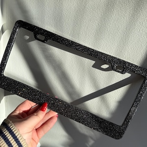May include: A black license plate frame with a glittery finish. The rectangular frame has rounded corners and mounting holes. The frame is held by a person with red nail polish.