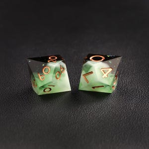 Op de afbeelding: Twee polyhedrale dobbelstenen met een groen en wit gemarmerd effect. De dobbelstenen hebben koperkleurige cijfers en randen. De bovenkant van elke dobbelsteen is zwart met glitter. De dobbelstenen zijn ontworpen voor tabletop games.