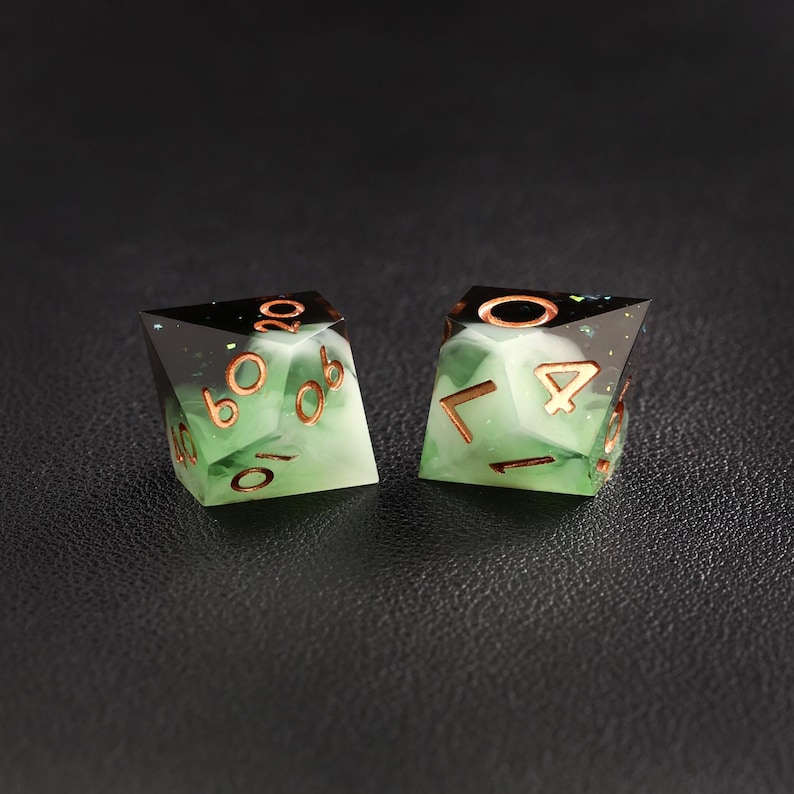 Op de afbeelding: Twee polyhedrale dobbelstenen met een groen en wit gemarmerd effect. De dobbelstenen hebben koperkleurige cijfers en randen. De bovenkant van elke dobbelsteen is zwart met glitter. De dobbelstenen zijn ontworpen voor tabletop games.