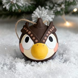 May include: A brown and white owl-shaped Christmas bauble with a yellow beak and pink cheeks. The decoration has a string for hanging and is set against a snowy background with a blurred Christmas tree.