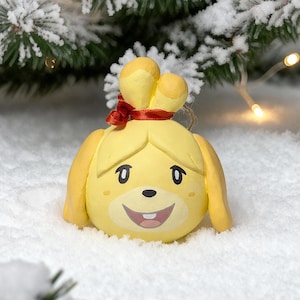 May include: A yellow, round, sculpted ornament featuring a character with long ears, a red ribbon tied at the top, and a smiling face with black eyes, a small nose, and a single visible tooth. The ornament has a smooth, matte finish.