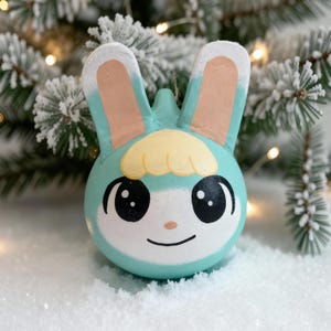 May include: A hand-painted, teal bunny ornament with large black eyes, a yellow tuft of hair, and pink-toned ears. The ornament is set against a snowy backdrop with a Christmas tree in the background. A string is attached for hanging.