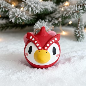 Peut inclure: Ornement de Noël en céramique rouge et blanche en forme de hibou, avec un bec jaune et des yeux noirs. L'ornement est suspendu à une ficelle, sur un fond enneigé avec un sapin de Noël flou.