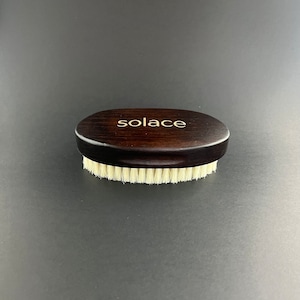 May include: A brown wooden brush with white bristles. The brush has the word "solace" engraved on the handle.