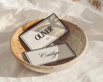 Pode incluir: Dois cartões de mesa com bordas pretas e os nomes "Oliver" e "Caitlyn" em fontes elegantes repousam em uma tigela de cerâmica salpicada. O cartão "Oliver" também tem "Table 2" impresso. A tigela está em uma superfície branca texturizada.