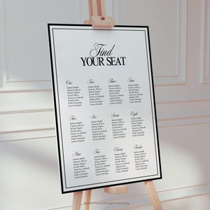 May include: A white wedding seating chart with a black border, displayed on a wooden easel. The chart features the text "Find Your Seat" in elegant script, with guest names organized by table numbers.