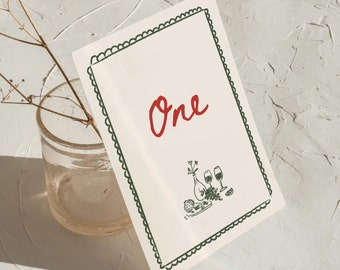 Könnte beinhalten: Eine cremefarbene Tischnummerkarte mit dem Wort "One" in roter Schrift. Die Karte hat einen grünen, gewellten Rand und eine Schwarz-Weiß-Illustration von Weingläsern und einer Flasche.