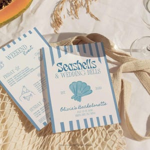 Könnte beinhalten: Zwei Einladungskarten mit blauen Streifen und Text. Eine Karte zeigt "Seashells & Wedding Bells" und "Olivia's Bachelorette" mit einer Muschelgrafik. Die andere Karte listet einen Wochenendveranstaltungsplan auf. Die Karten liegen auf einer beigen Netztasche.