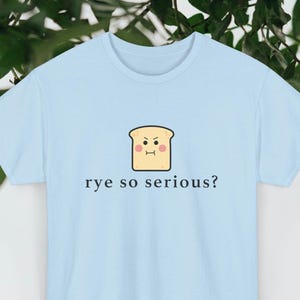 May include: Pale blue t-shirt featuring the phrase "rye so serious?" and a cartoon bread slice with a displeased expression. The t-shirt is a light blue colour.
