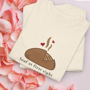 Puede incluir: Dos camisetas color crema apiladas, con un gr&aacute;fico de una hogaza de pan marr&oacute;n con vapor y corazones. El texto "loaf at first sight" est&aacute; debajo del pan. La palabra "Natural" est&aacute; en un cuadro blanco en la esquina superior derecha.