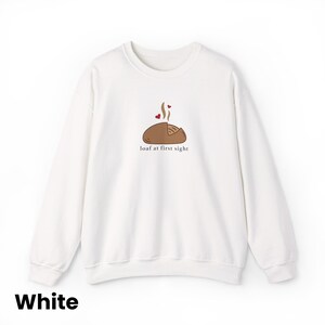 Peut inclure: Sweat-shirt blanc &agrave; col rond avec un motif de pain. Le motif comprend un pain de dessin anim&eacute; avec de la vapeur et des c&oelig;urs, et la phrase "loaf at first sight". Un style confortable et d&eacute;contract&eacute;.