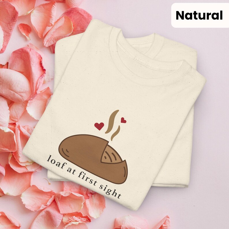 Puede incluir: Dos camisetas color crema apiladas, con un gr&aacute;fico de una hogaza de pan marr&oacute;n con vapor y corazones. El texto "loaf at first sight" est&aacute; debajo del pan. La palabra "Natural" est&aacute; en un cuadro blanco en la esquina superior derecha.