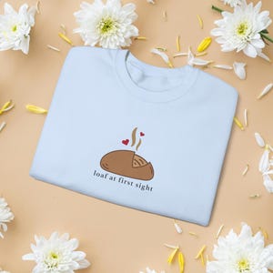 Peut inclure: Un sweat-shirt bleu clair avec un dessin de pain de dessin anim&eacute;, de la vapeur et deux c&oelig;urs. Le texte "loaf at first sight" est en dessous de l'illustration du pain. Des fleurs et des p&eacute;tales blancs sont dispers&eacute;s autour du sweat-shirt.