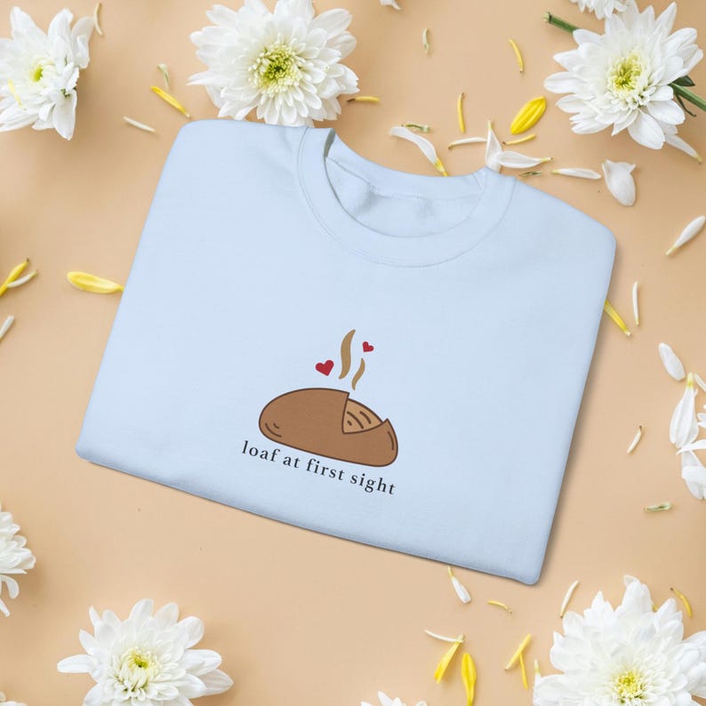 Peut inclure: Un sweat-shirt bleu clair avec un dessin de pain de dessin anim&eacute;, de la vapeur et deux c&oelig;urs. Le texte "loaf at first sight" est en dessous de l'illustration du pain. Des fleurs et des p&eacute;tales blancs sont dispers&eacute;s autour du sweat-shirt.