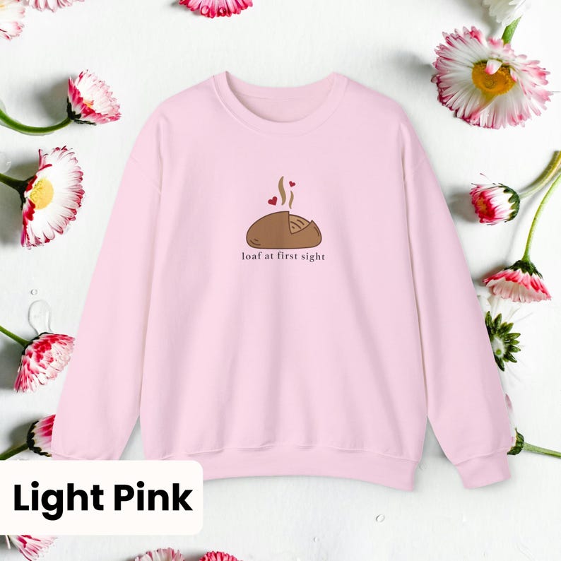 Peut inclure: Sweat-shirt rose clair avec un dessin d'un pain avec de la vapeur et des coeurs, et le texte "loaf at first sight". Le sweat-shirt est entour&eacute; de fleurs blanches et roses.