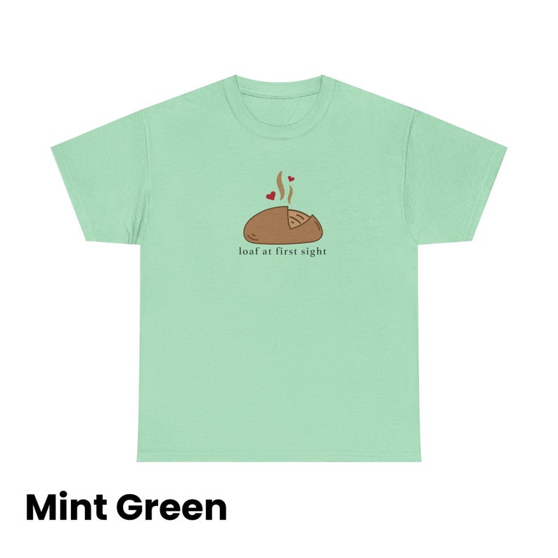 Puede incluir: Camiseta verde menta con un gr&aacute;fico de una hogaza de pan con vapor y dos corazones, y el texto "loaf at first sight". La camiseta es de color verde menta liso.