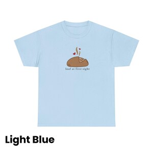 Puede incluir: Camiseta azul claro con un gr&aacute;fico de una hogaza de pan con vapor y corazones, y el texto "loaf at first sight". La camiseta es de color azul claro.