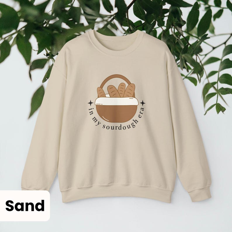 Peut inclure: Sweat-shirt couleur sable avec un motif de panier &agrave; pain et le texte "in my sourdough era". L'illustration pr&eacute;sente un panier marron avec des miches de pain. Le sweat-shirt a un col rond et des manches longues.