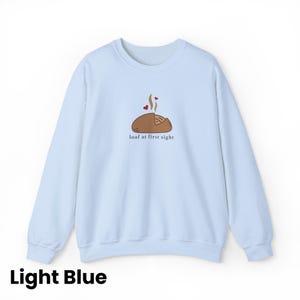Peut inclure: Sweat-shirt bleu clair avec un dessin d'un pain avec de la vapeur et des coeurs, et le texte "loaf at first sight". Le sweat-shirt est de couleur bleu clair uni.