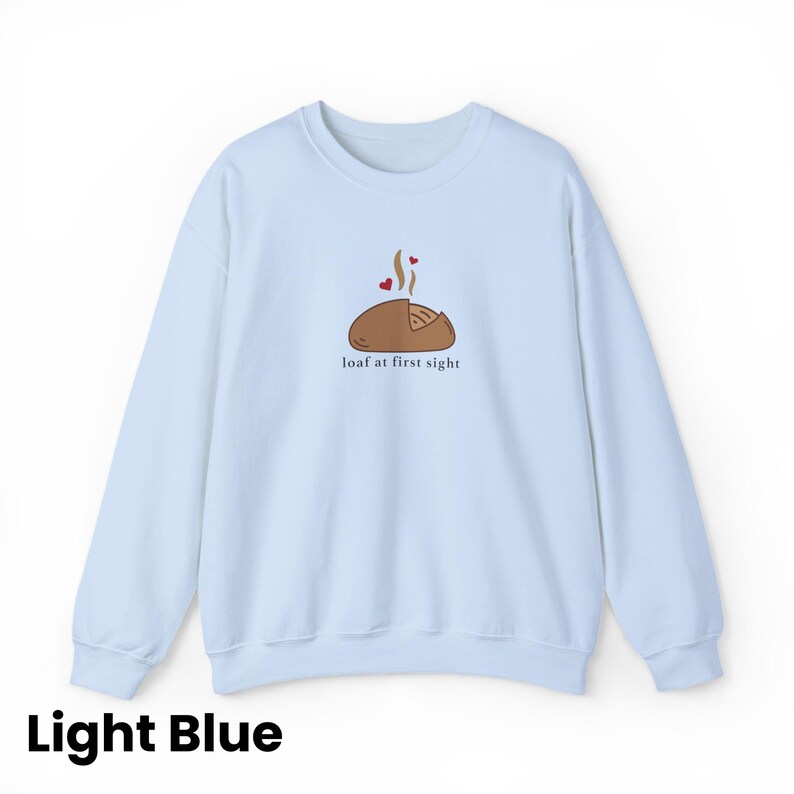 Peut inclure: Sweat-shirt bleu clair avec un dessin d'un pain avec de la vapeur et des coeurs, et le texte "loaf at first sight". Le sweat-shirt est de couleur bleu clair uni.
