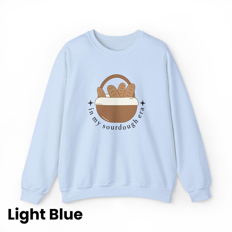 Peut inclure: Sweat-shirt bleu clair &agrave; col rond. Il pr&eacute;sente un motif de panier marron rempli de pains, avec le texte "in my sourdough era" dans un design circulaire. Les mots "Light Blue" sont en bas.
