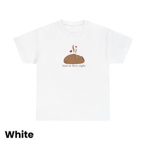Puede incluir: Camiseta blanca con un gr&aacute;fico de pan marr&oacute;n, vapor y dos corazones rojos. El texto "loaf at first sight" est&aacute; debajo de la ilustraci&oacute;n del pan. La palabra "White" est&aacute; en la parte inferior de la camiseta.