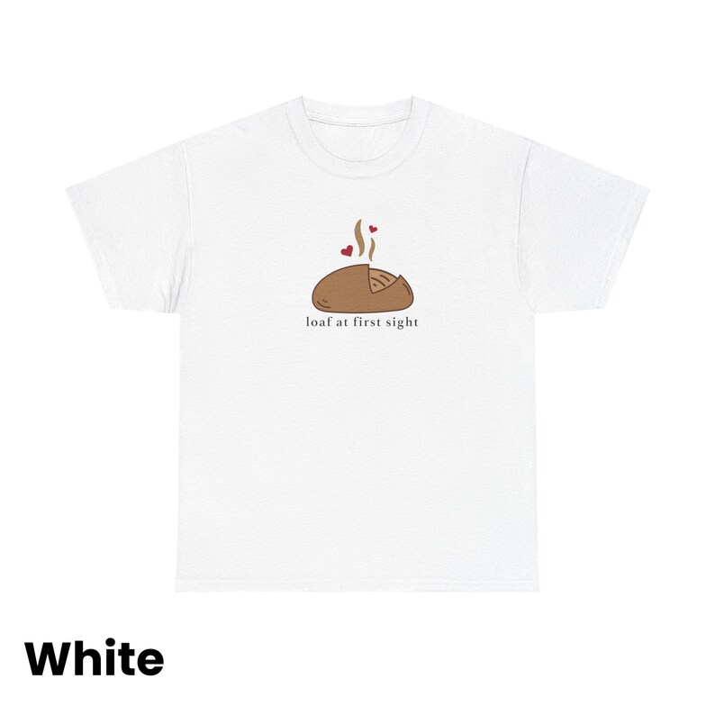 Puede incluir: Camiseta blanca con un gr&aacute;fico de pan marr&oacute;n, vapor y dos corazones rojos. El texto "loaf at first sight" est&aacute; debajo de la ilustraci&oacute;n del pan. La palabra "White" est&aacute; en la parte inferior de la camiseta.