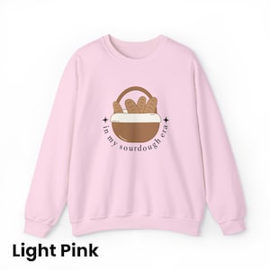 Peut inclure: Sweat-shirt rose clair avec un motif de panier &agrave; pain et le texte "in my sourdough era". Le motif pr&eacute;sente un panier marron avec des miches de pain et le texte en cercle. Le sweat-shirt est de couleur rose clair.