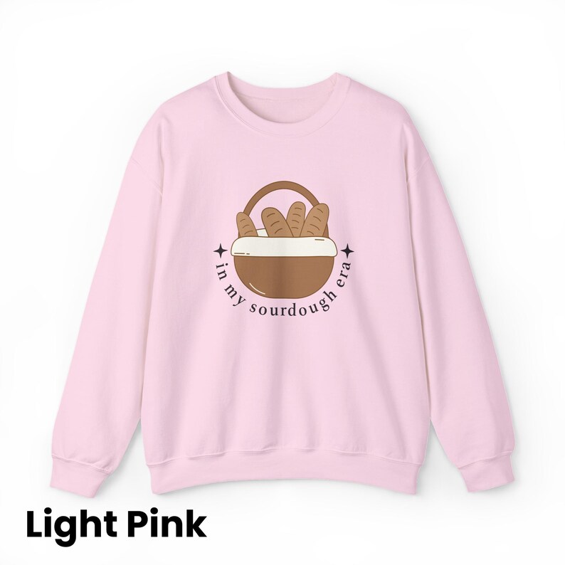 Peut inclure: Sweat-shirt rose clair avec un motif de panier &agrave; pain et le texte "in my sourdough era". Le motif pr&eacute;sente un panier marron avec des miches de pain et le texte en cercle. Le sweat-shirt est de couleur rose clair.