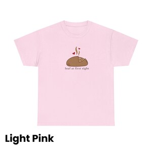 Puede incluir: Camiseta rosa claro con un gr&aacute;fico de una hogaza de pan con vapor y corazones, y el texto "loaf at first sight". La camiseta es de color rosa claro.